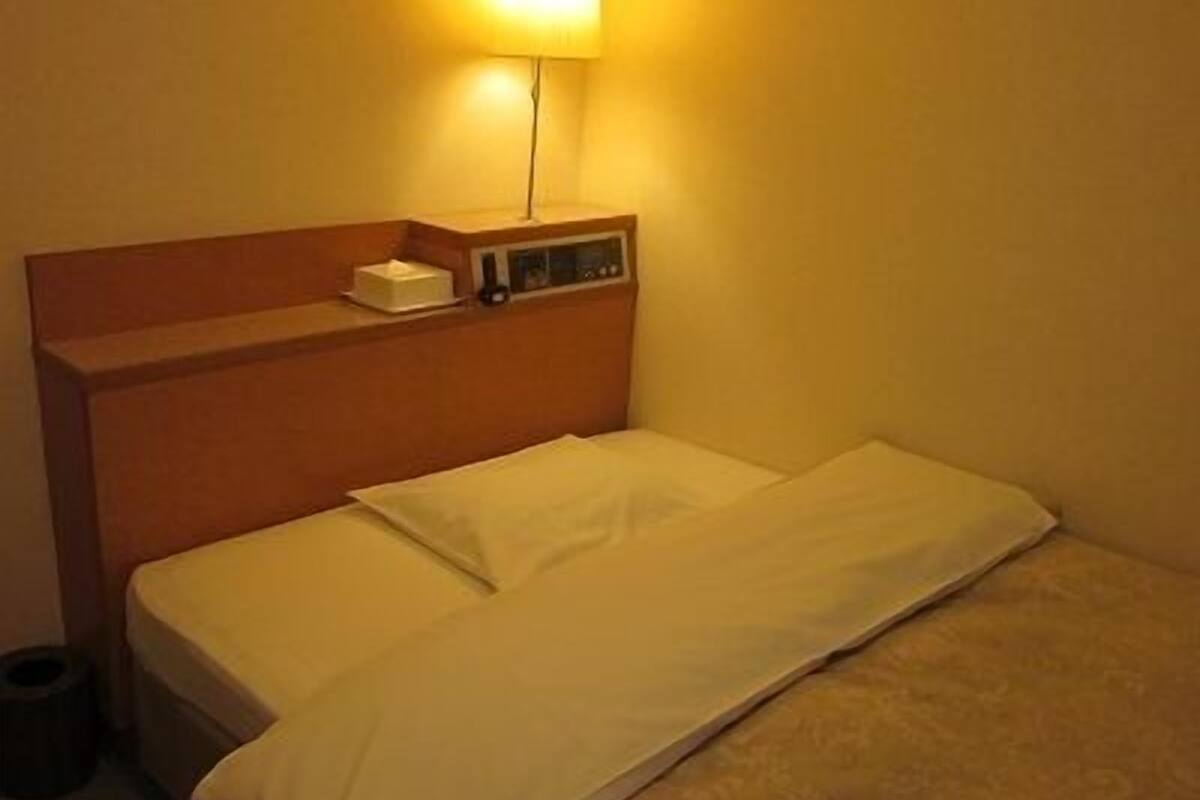 A single bed with a wooden headboard, a bedside table with a lamp, and a small shelf with a box and a remote control.