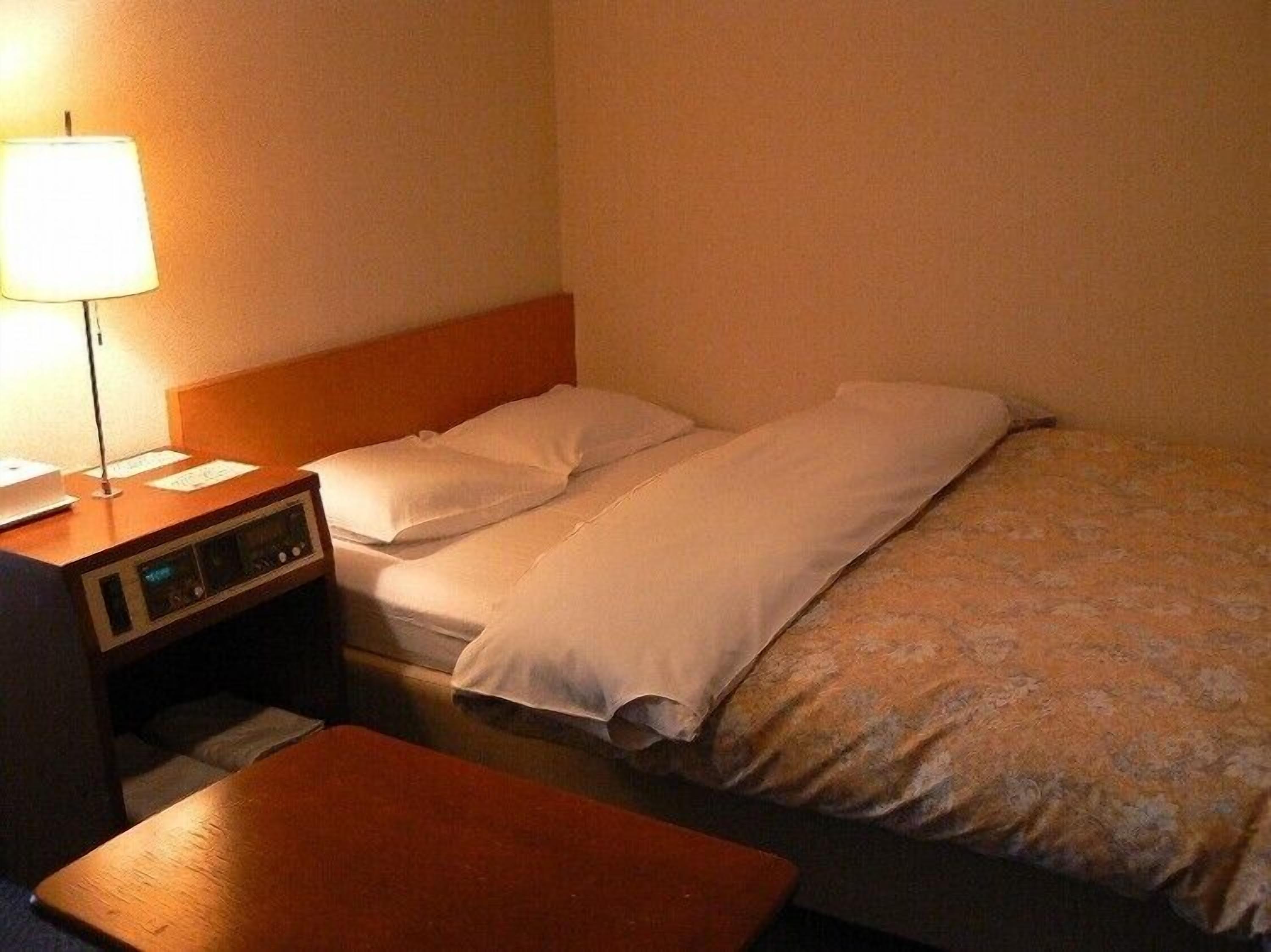 A hotel room with a bed, a bedside table with a lamp, and a small desk with a computer.