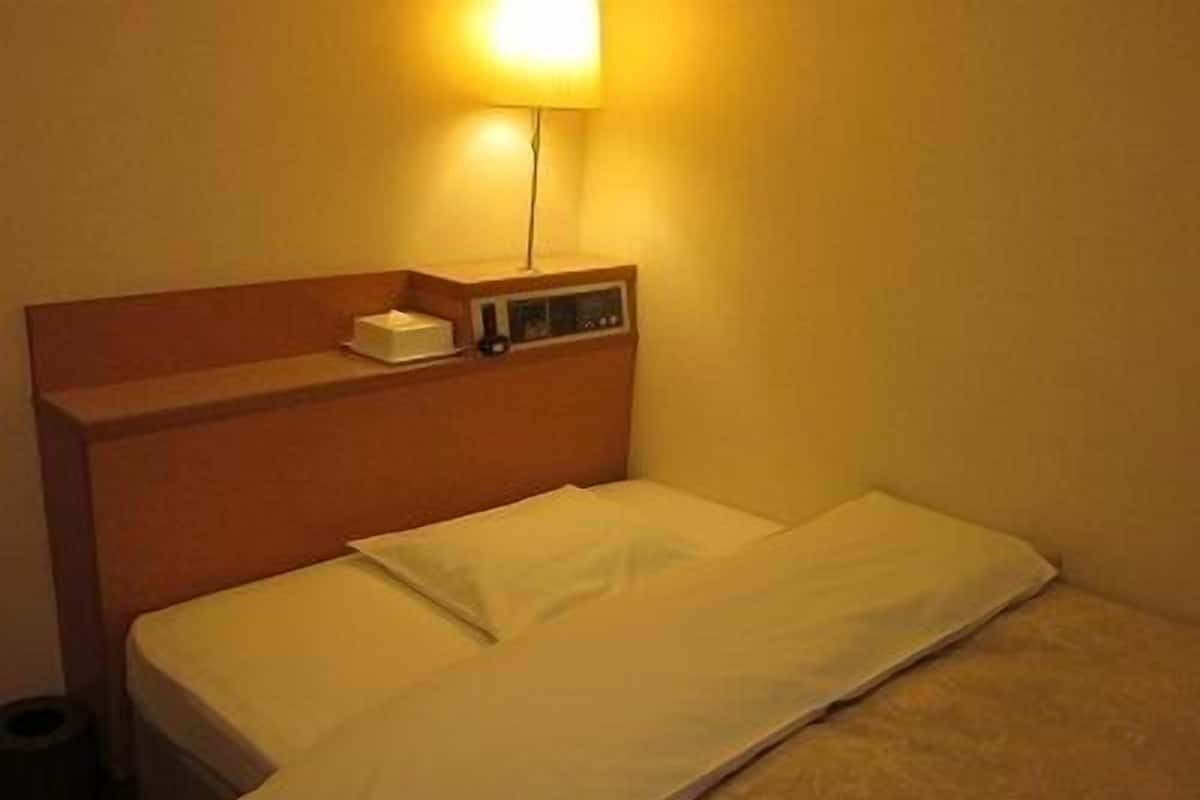 A single bed with a wooden headboard, a bedside table with a lamp, and a small shelf with a box and a remote control.