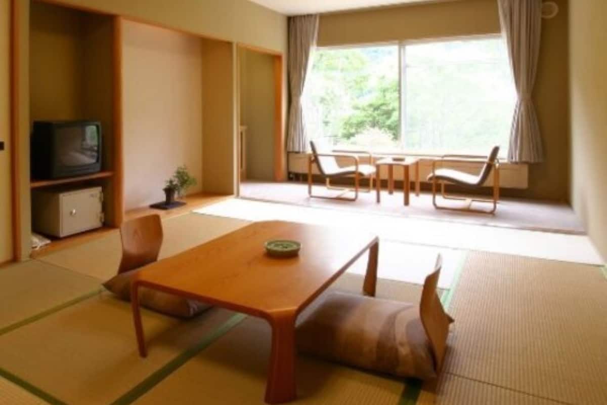 Ein Zimmer mit Tatami-Boden, einem Holztisch, einem Fernseher und einem Fenster mit Vorhängen.