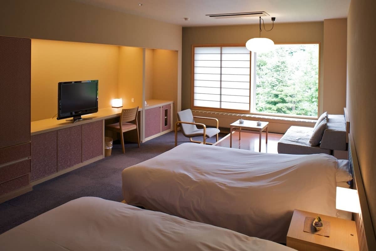 Ein Hotelzimmer mit zwei Betten, einem Schreibtisch mit Fernseher, einem Sessel, einem kleinen Tisch und einem Fenster mit Blick ins Grüne.