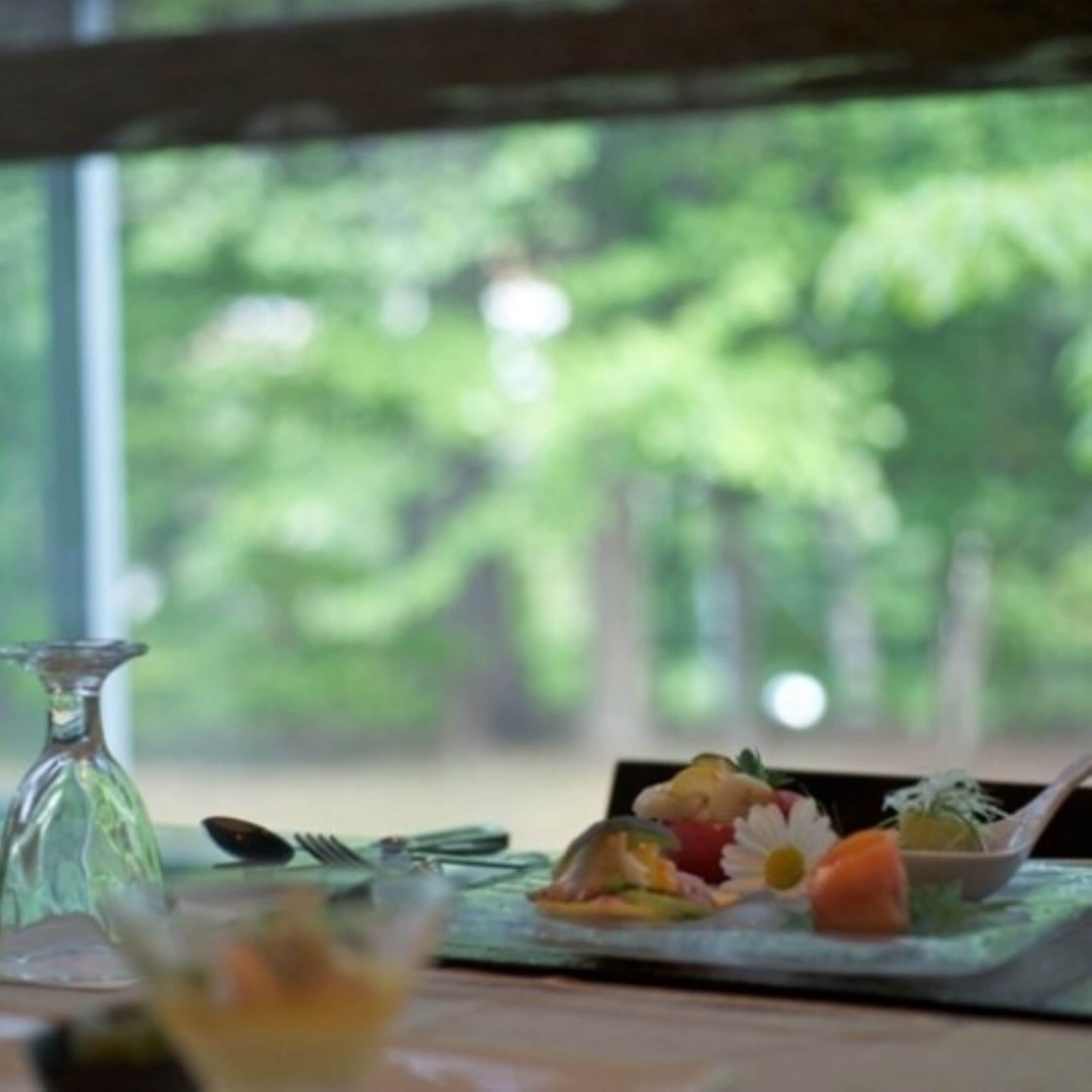 Una mesa de comedor con comida servida, una botella de vidrio y vistas al exterior con vegetación.
