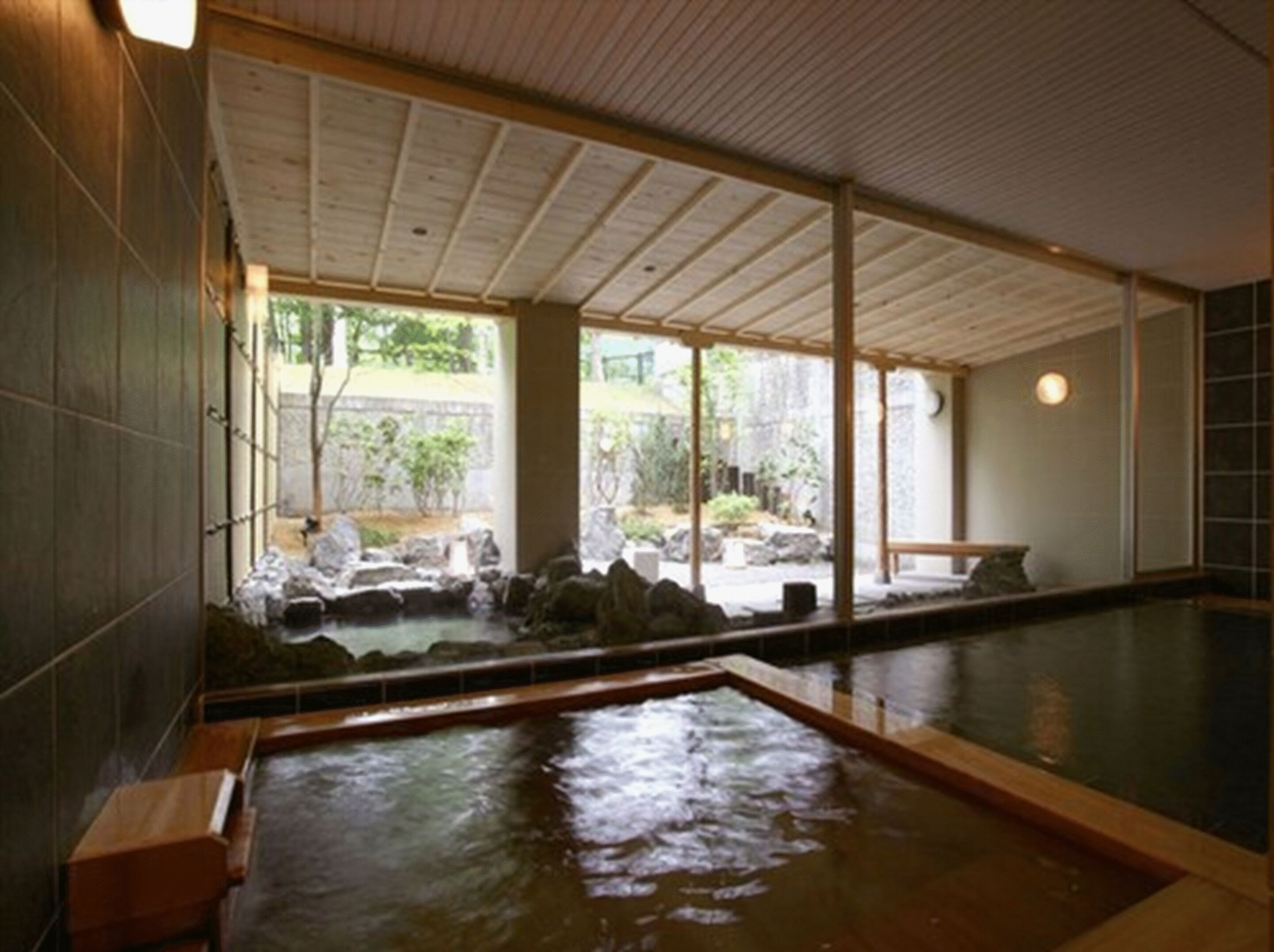 A traditional Japanese onsen with a wooden interior, a stone bath, and a view of a garden through large windows.