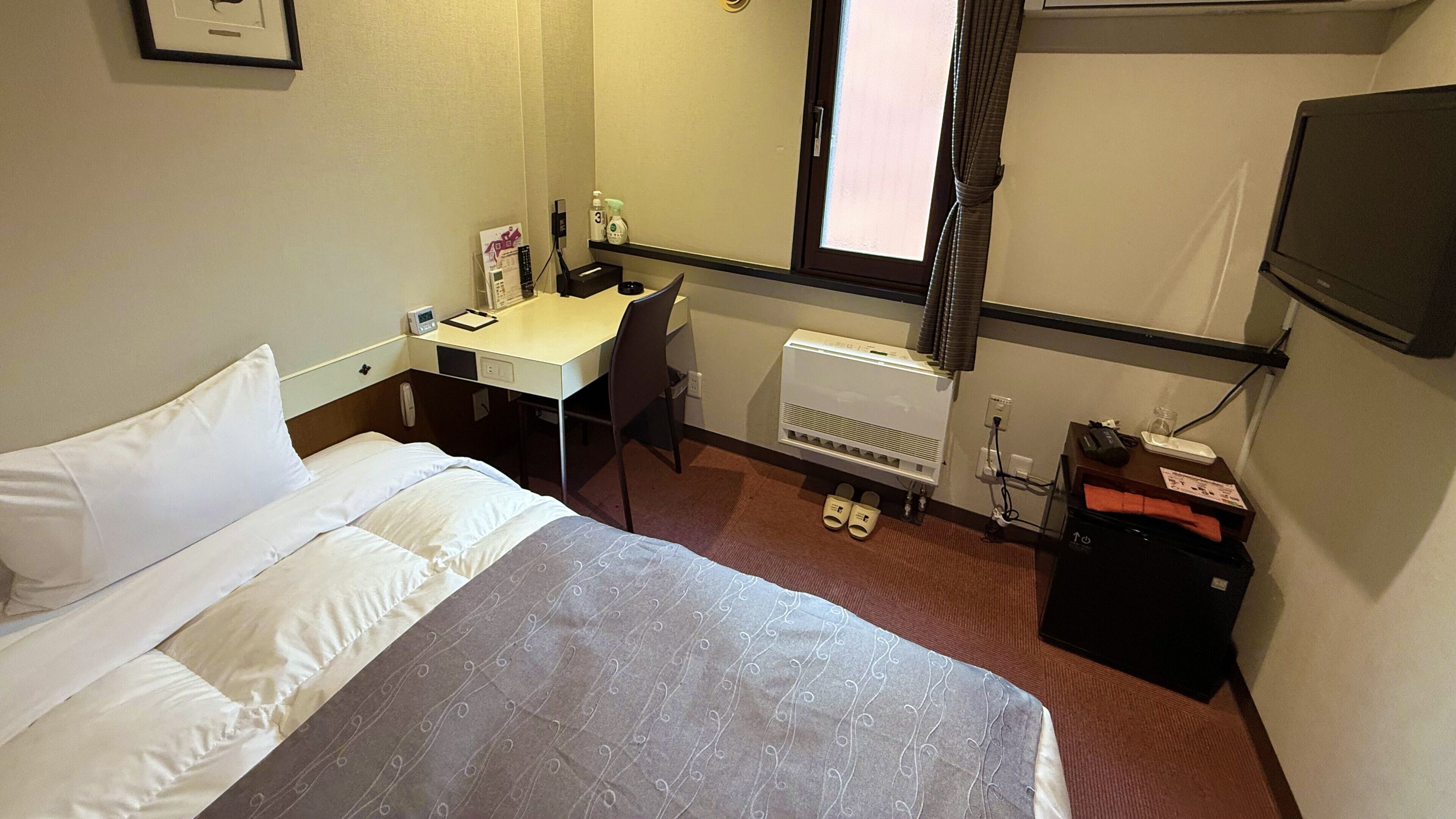 A hotel room with a bed, desk, chair, window, radiator, and a TV.