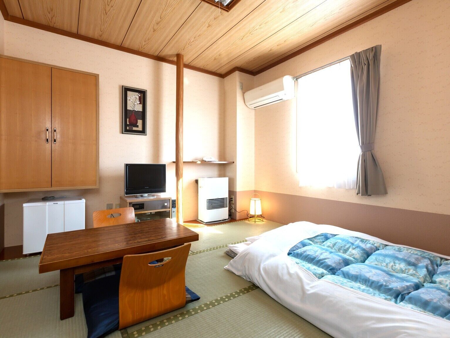 Habitación de hotel con techo de madera, cama con cubrecama azul y blanco, televisor sobre un mueble de madera, aire acondicionado blanco y un armario de madera.