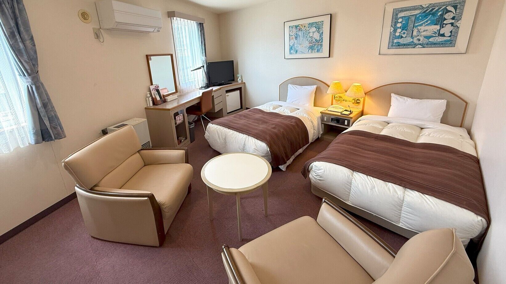 Habitación de hotel con dos camas, un escritorio, una silla, una mesita y obras de arte en las paredes.
