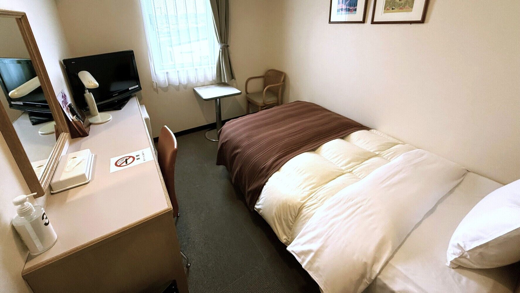 Habitación de hotel con una cama, un escritorio con computadora, una silla, una mesita y una ventana con cortinas.