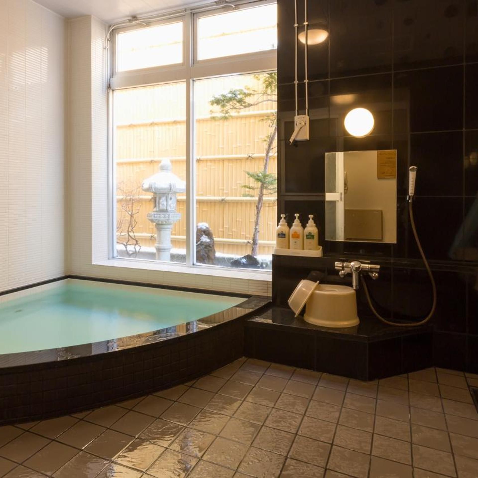 A spa room with a large window, a hot tub, a shower area, and a small fountain outside.