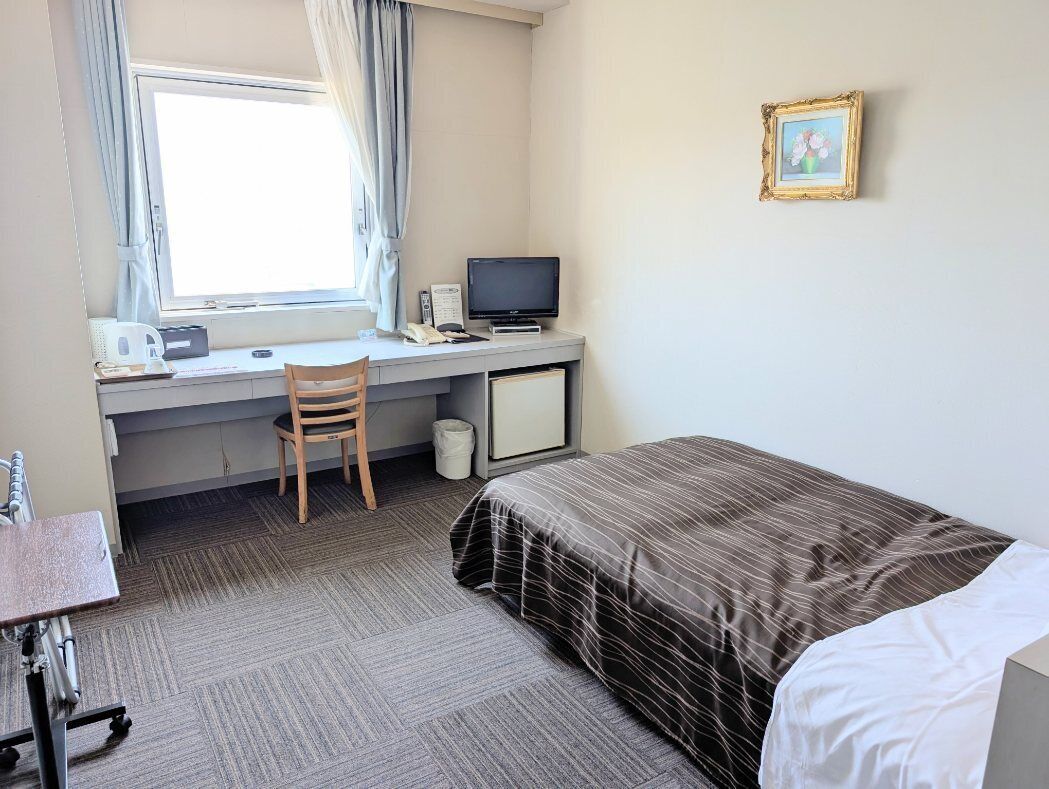 A hotel room with a bed, a desk with a computer, a chair, and a window with curtains.
