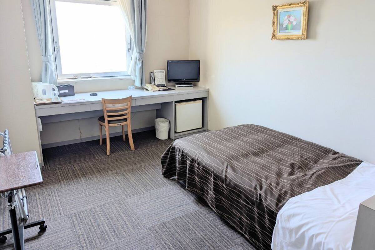 A hotel room with a bed, a desk with a computer, a chair, and a window with curtains.