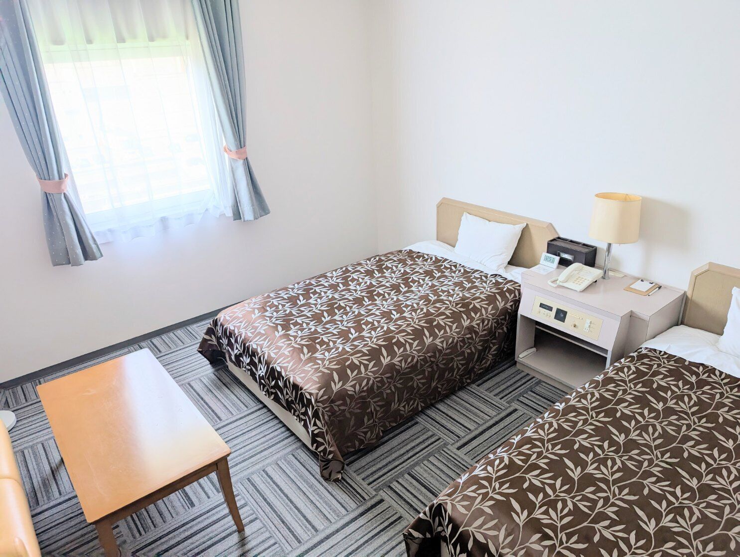 A hotel room with a bed, bedside table, lamp, and a small desk with a telephone.