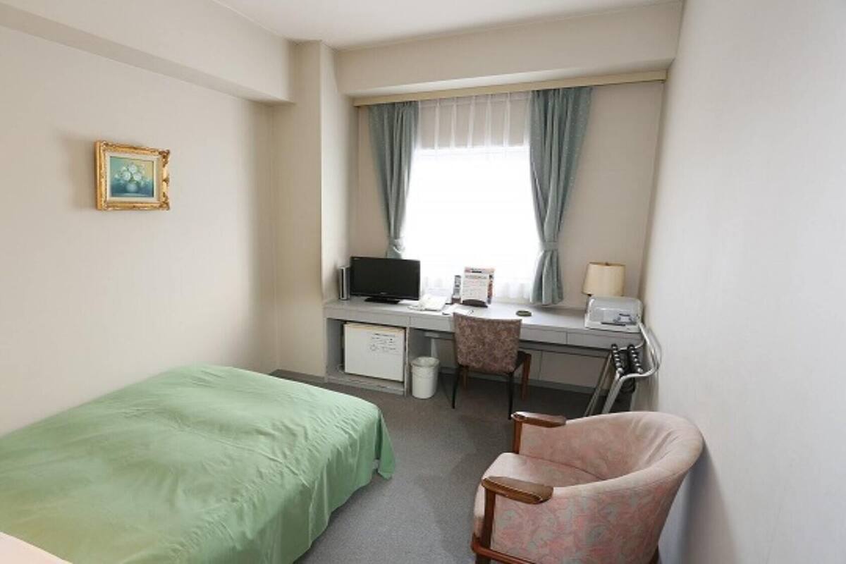 A hotel room with a single bed, a desk with a computer, a chair, and a window with curtains.