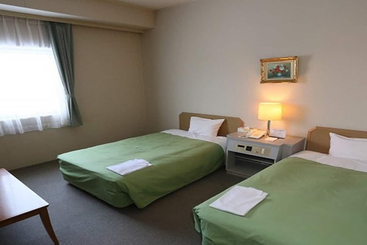 A hotel room with two single beds, a nightstand with a lamp, a small table, and a window with curtains.
