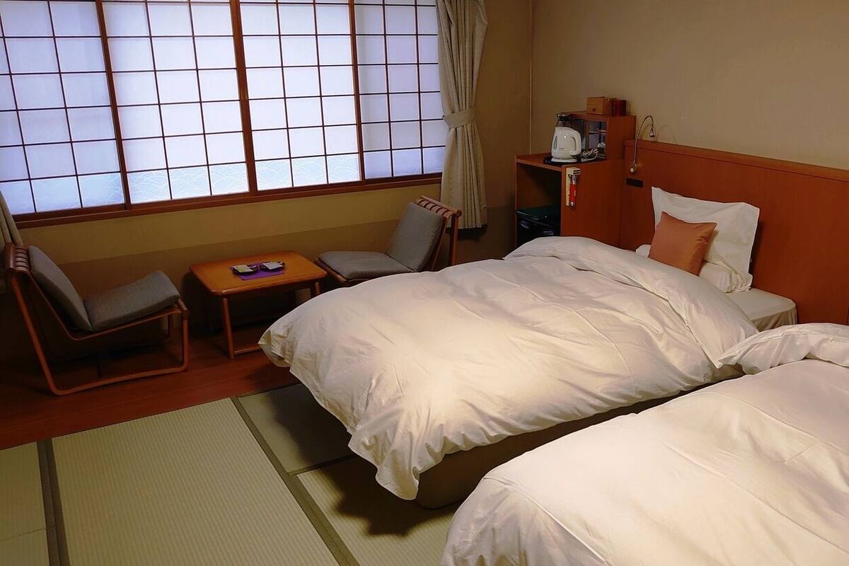 A hotel room with two beds, a small table, a chair, and a window with a view.