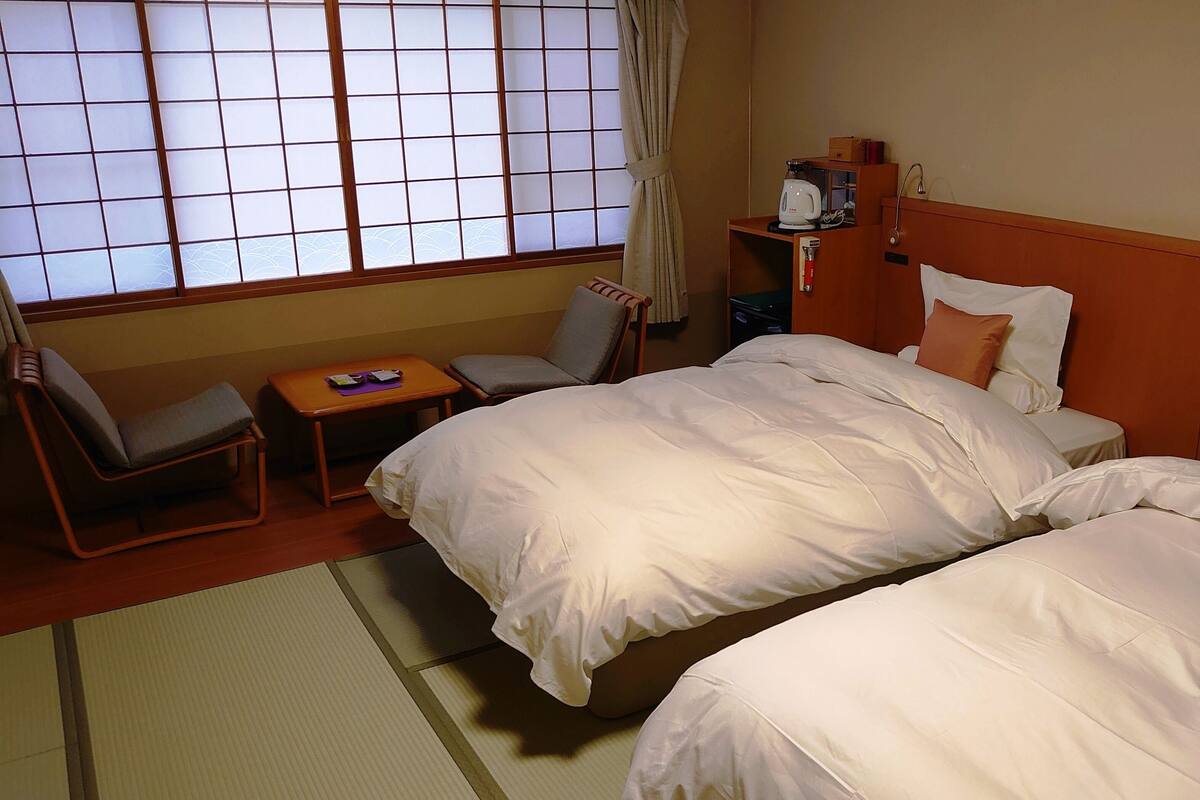 A hotel room with a bed, two chairs, a small table, a window with curtains, and a shelf with a kettle.