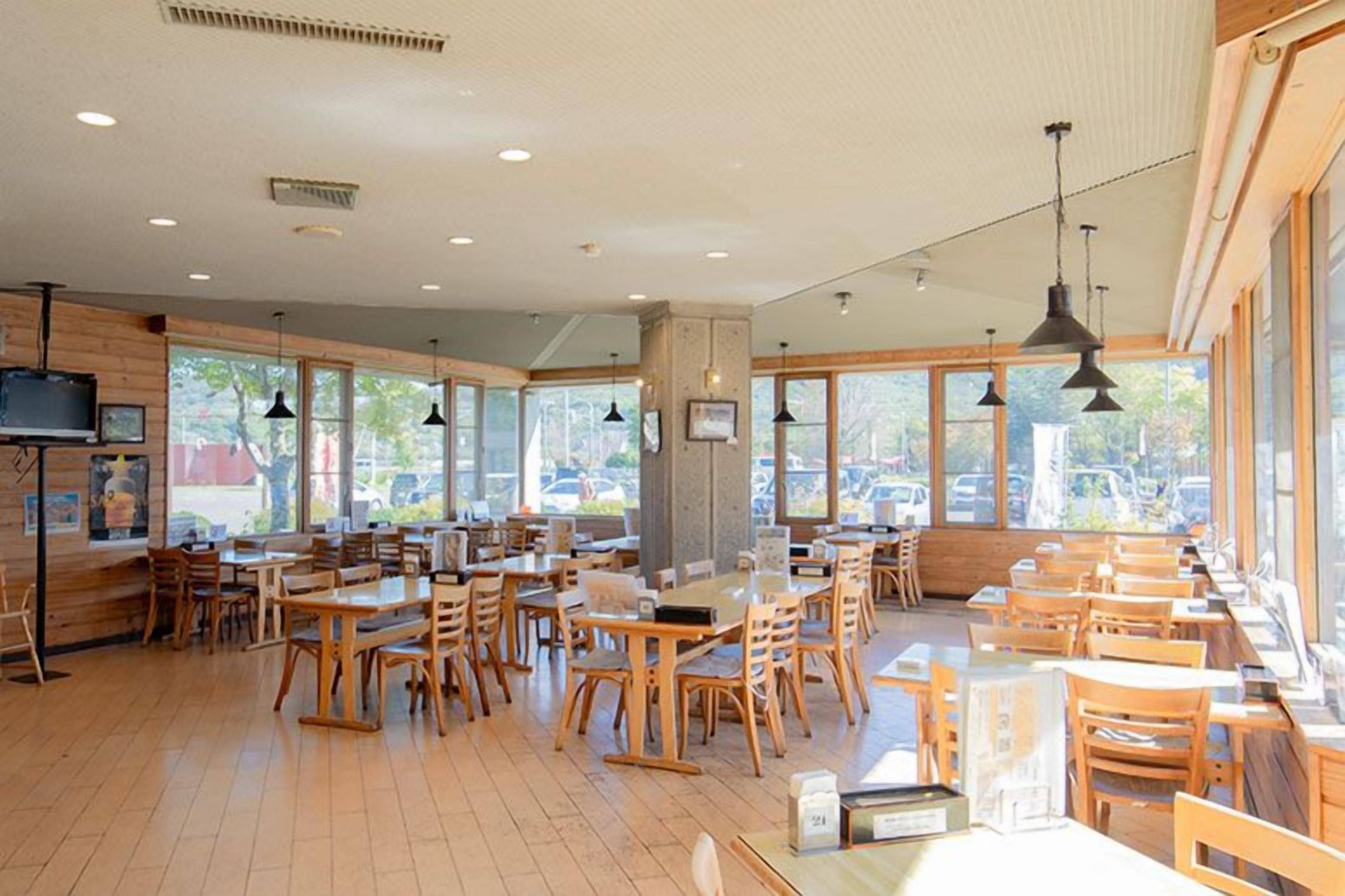 Un restaurante espacioso con mesas y sillas de madera, ventanas amplias e iluminación colgante.