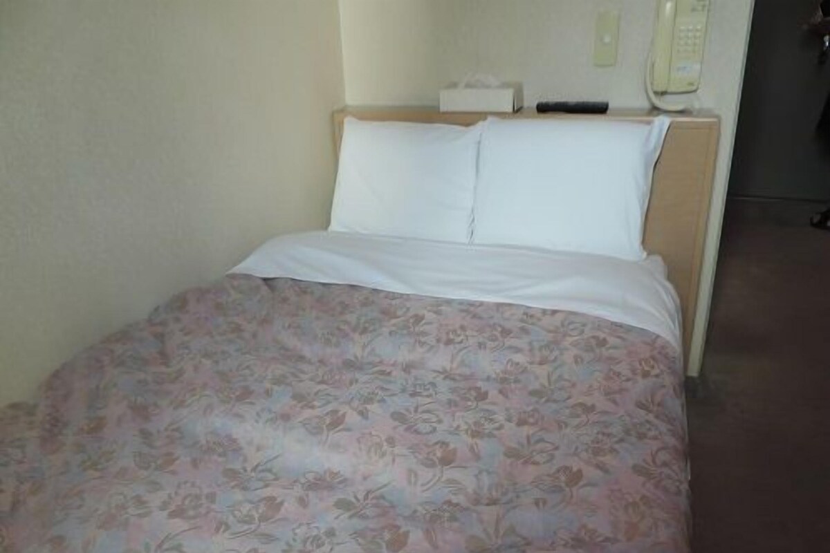 A bed with a floral bedspread, a white pillow, and a bedside table with a tissue box and a phone.