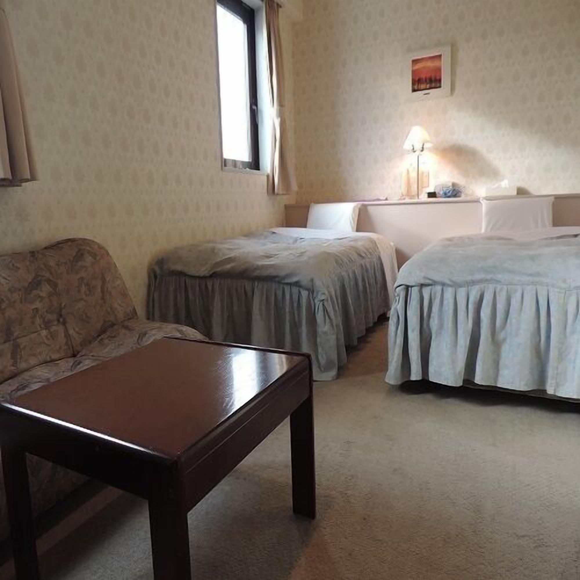 Ein Hotelzimmer mit einer Couch, zwei Einzelbetten, einem Holztisch, einem Fenster und einer Wandleuchte.
