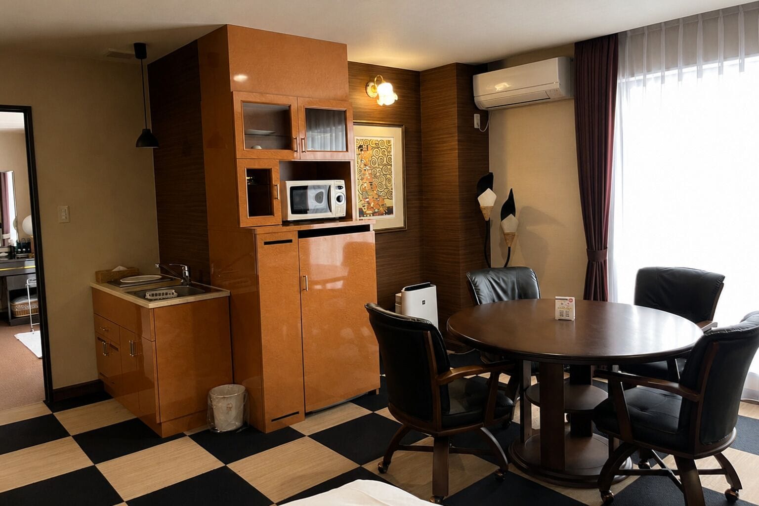 Une chambre d’hôtel compacte dotée d’une kitchenette, d’un coin repas avec une table ronde et des chaises, d’une poubelle et d’une grande fenêtre avec des rideaux.