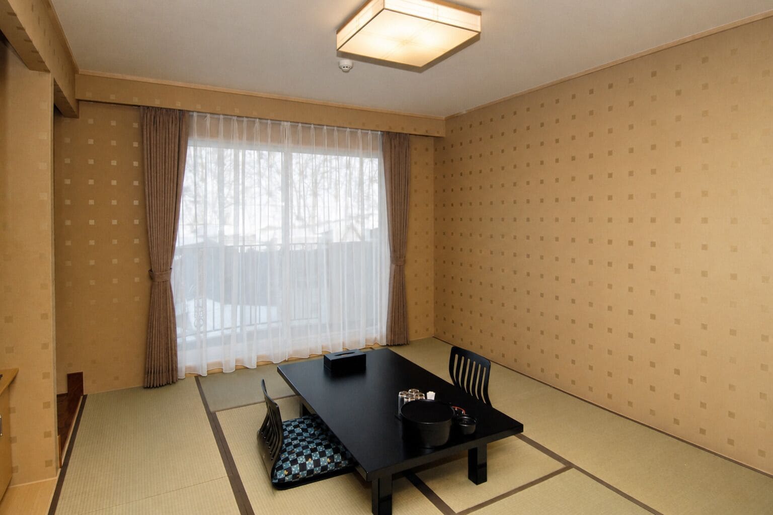A room with tatami flooring, a black table with chairs, a window with sheer curtains, and a patterned wall.