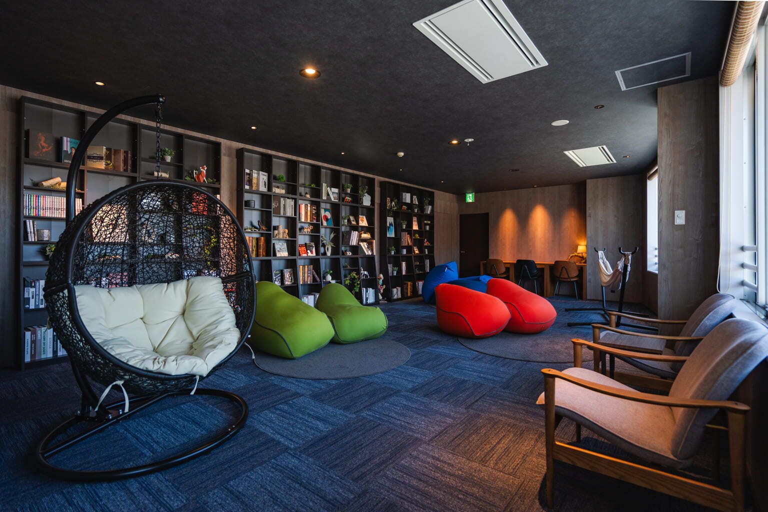 A modern lounge area with a large bookshelf, a hanging chair, and colorful bean bags.