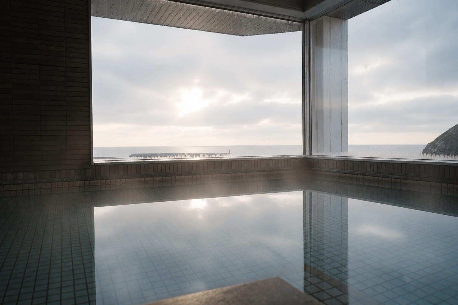A modern indoor swimming pool with a clear view of the sea and a distant pier.