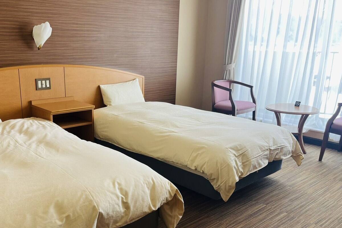 A hotel room with two beds, a nightstand, a chair, a small table, and a window with curtains.