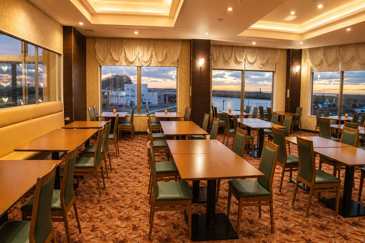 A hotel dining area with multiple tables and chairs, large windows with curtains, and a carpeted floor.