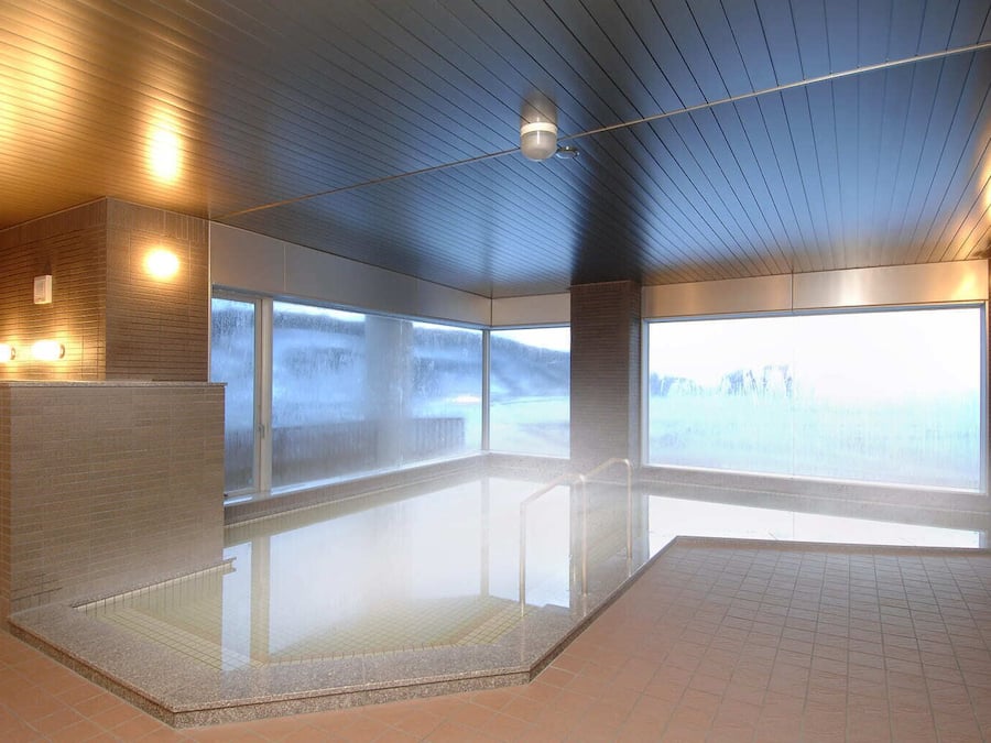 Un área de piscina interior moderna con paredes de vidrio transparente, piso de baldosas e iluminación lineal en el techo.