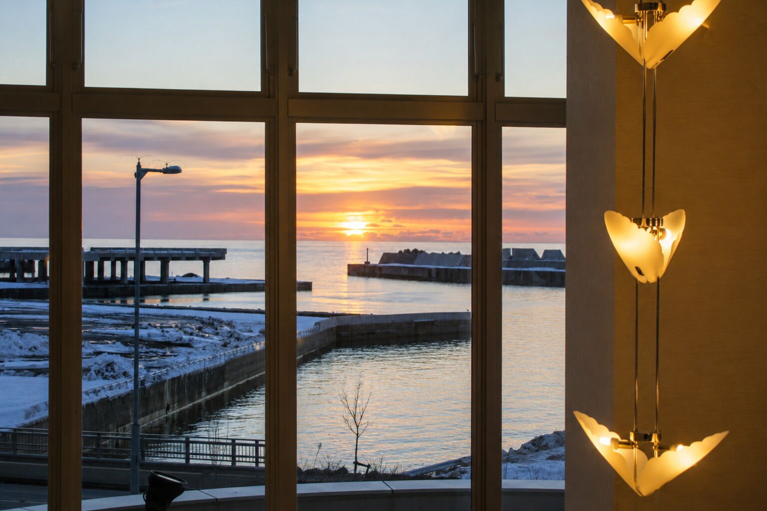 A view through a window with a sunset over a body of water, a pier, and a snow-covered landscape.
