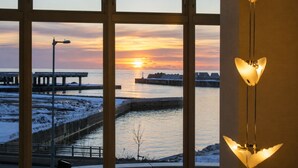 Udsigt gennem et vindue med solnedgang over vand, en mole og et snedækket landskab.