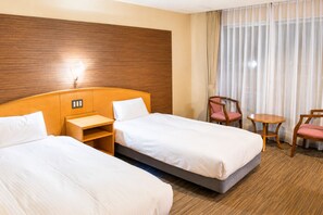 Habitación de hotel con dos camas, cabecero de madera, una mesita, dos sillas y una ventana con cortinas.