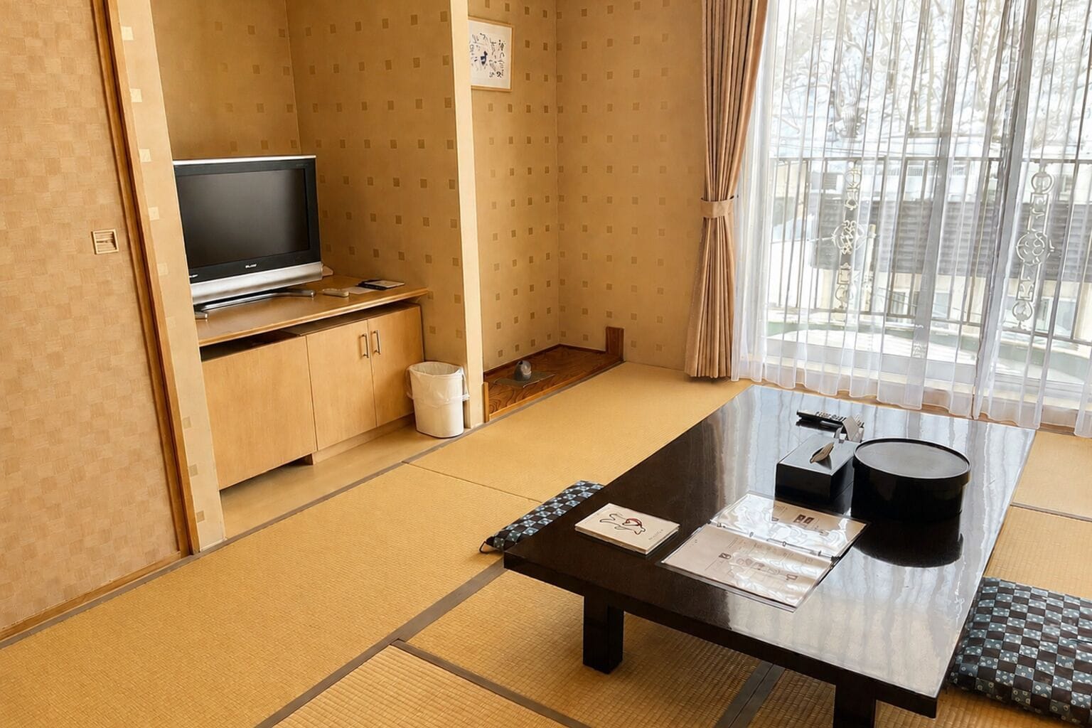 A room with tatami flooring, a television, a desk with papers and a black container, and a window with curtains.