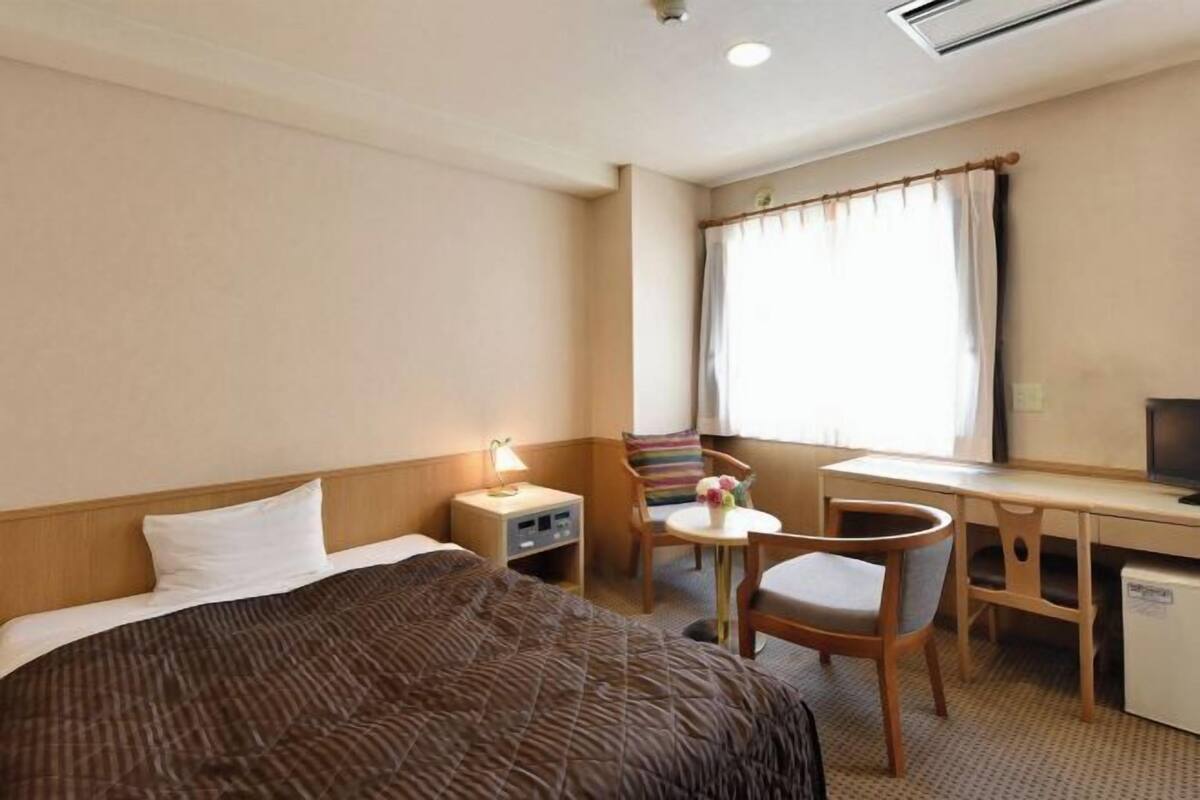 A hotel room with a bed, a desk, a chair, a small table, and a window with curtains.