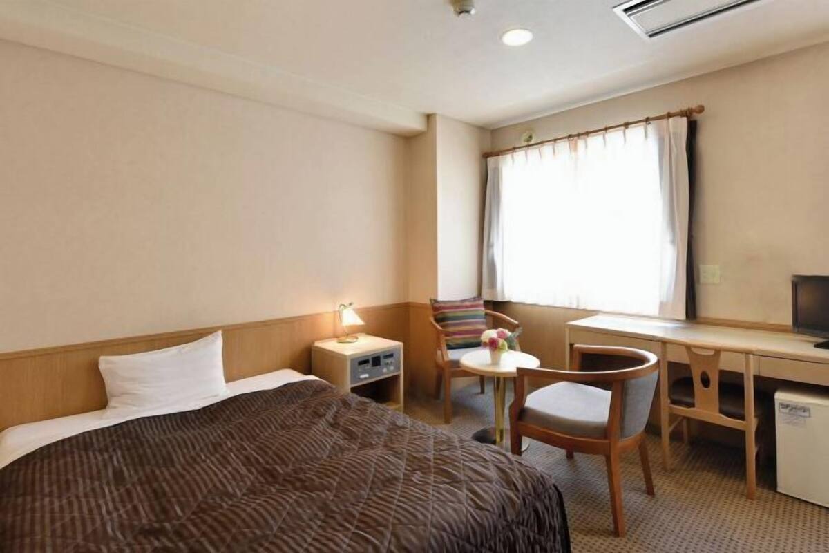 A hotel room with a bed, a desk, a chair, a small table, a lamp, and a window with curtains.