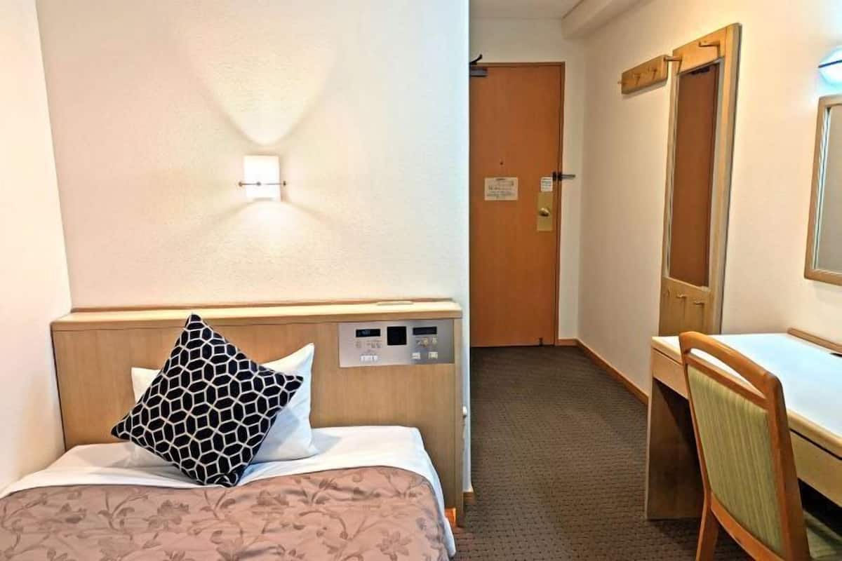 A hotel room with a bed, a desk, a chair, a wall-mounted control panel, and a door.