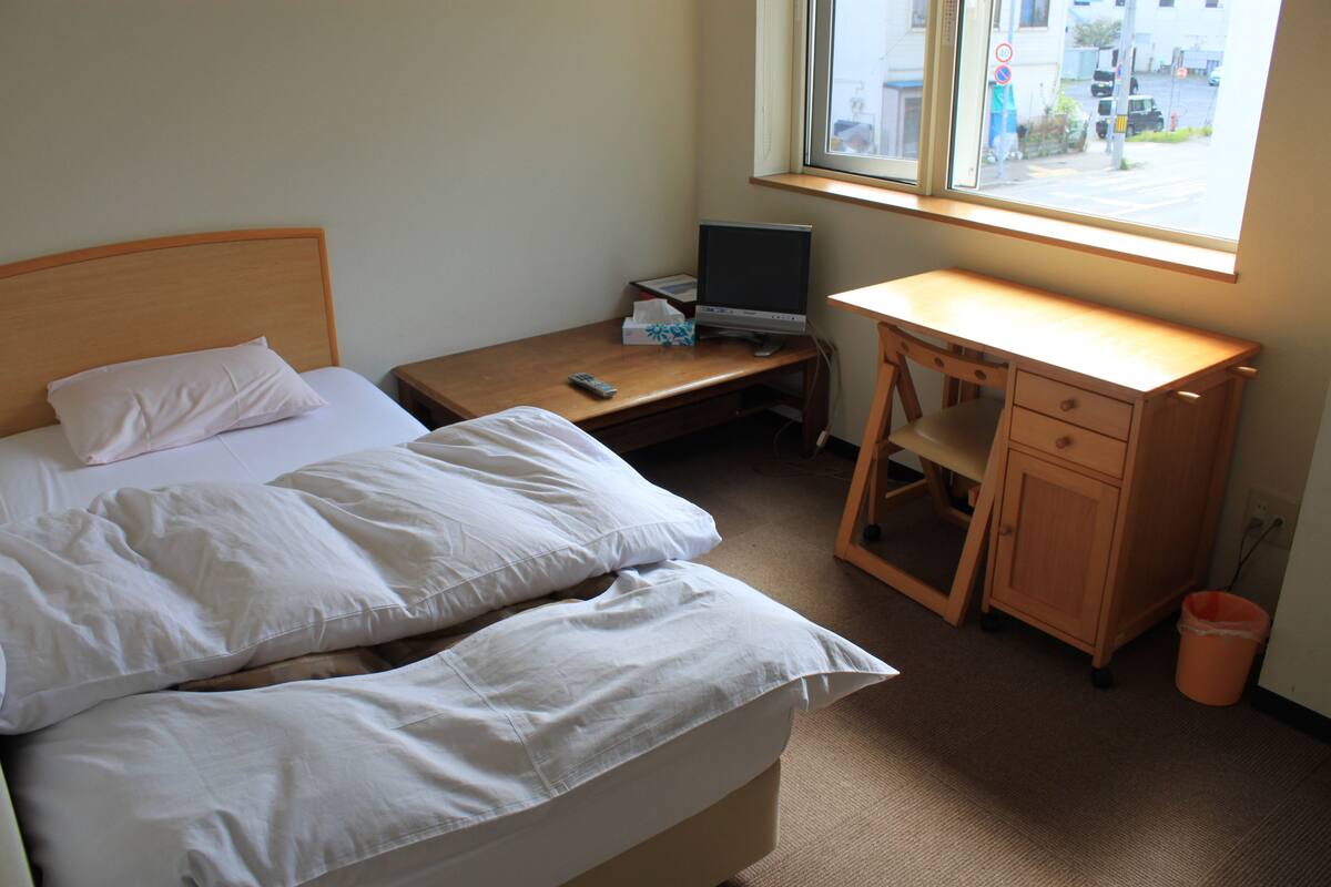 A bedroom with a bed, a desk, a TV, and a window.