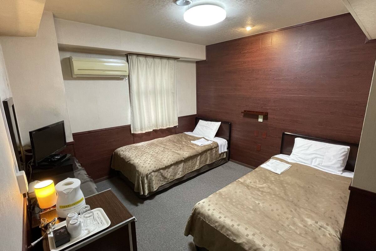 A hotel room with two beds, a desk with a lamp, a TV, and an air conditioning unit.