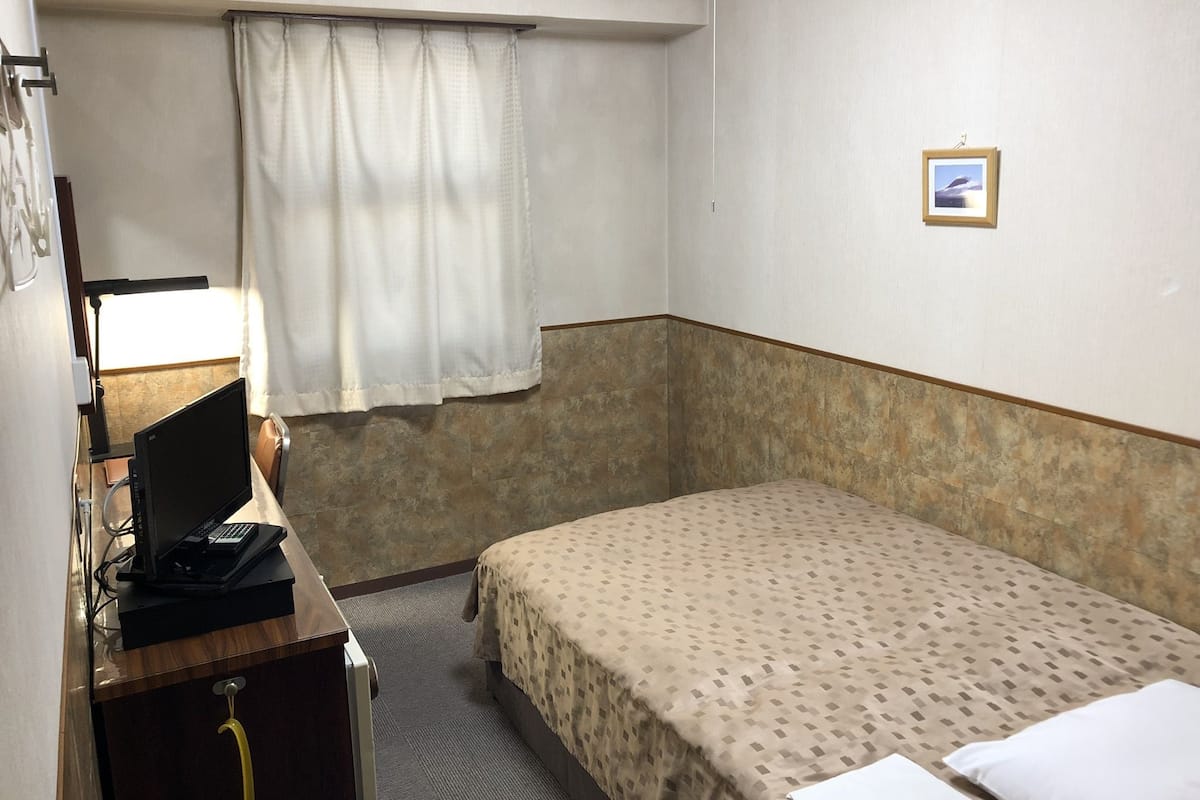 A small hotel room with a single bed, a desk with a computer, a window with curtains, and a small picture on the wall.