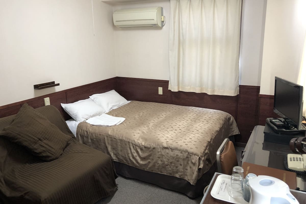 A hotel room with a bed, a desk with a computer, a chair, a small table with a phone and a glass, and a window with curtains.