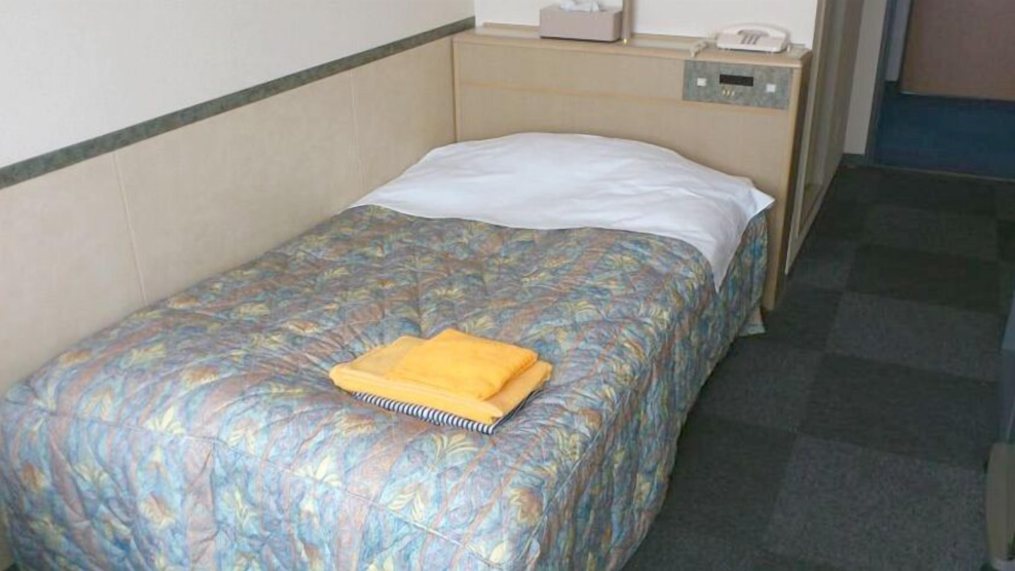 A single bed with a patterned bedspread, a white pillow, and a folded yellow towel on the bed.
