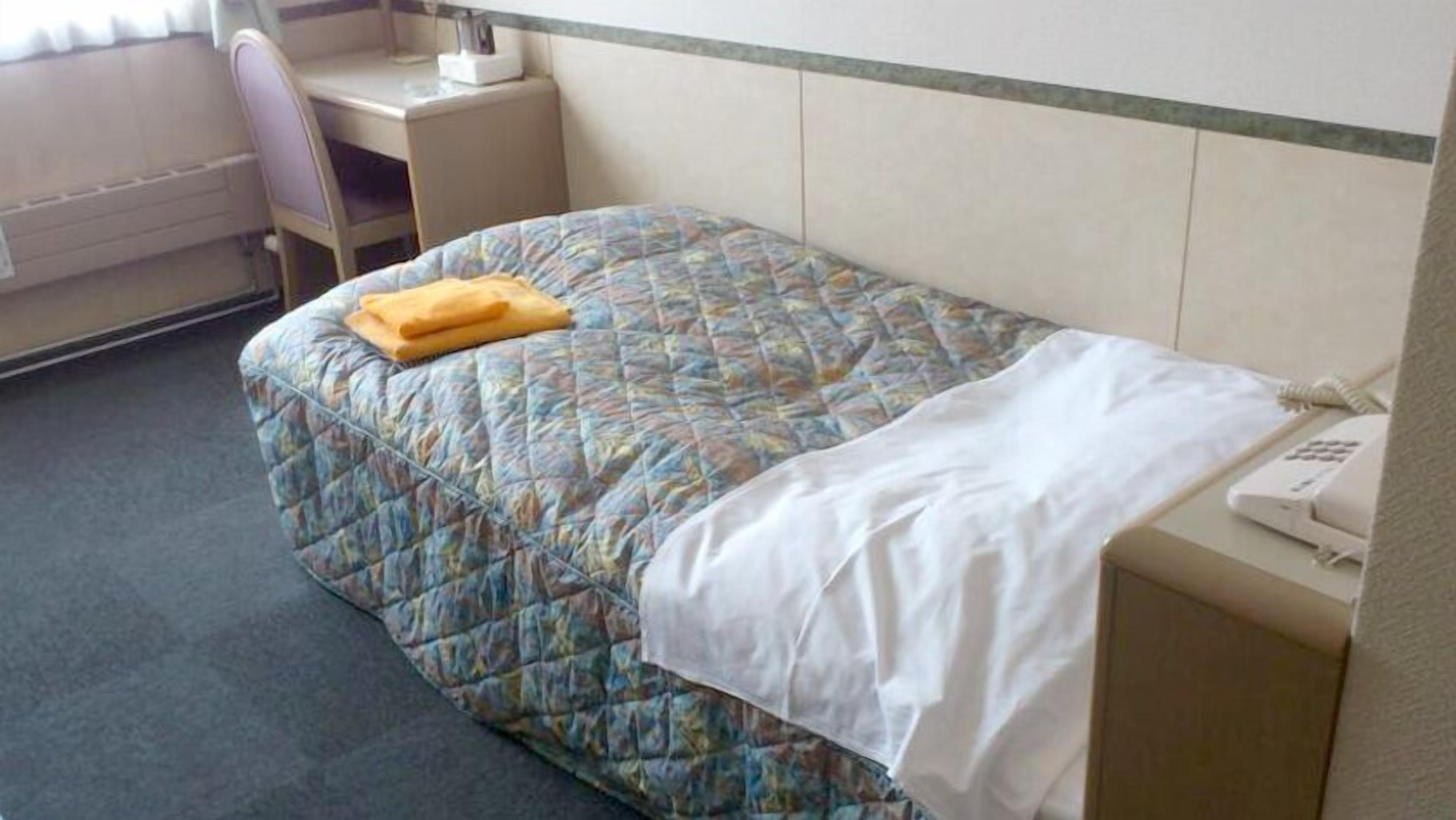 A single bed with a patterned bedspread, a white blanket, and a yellow towel. There is a desk with a chair and a telephone on it.