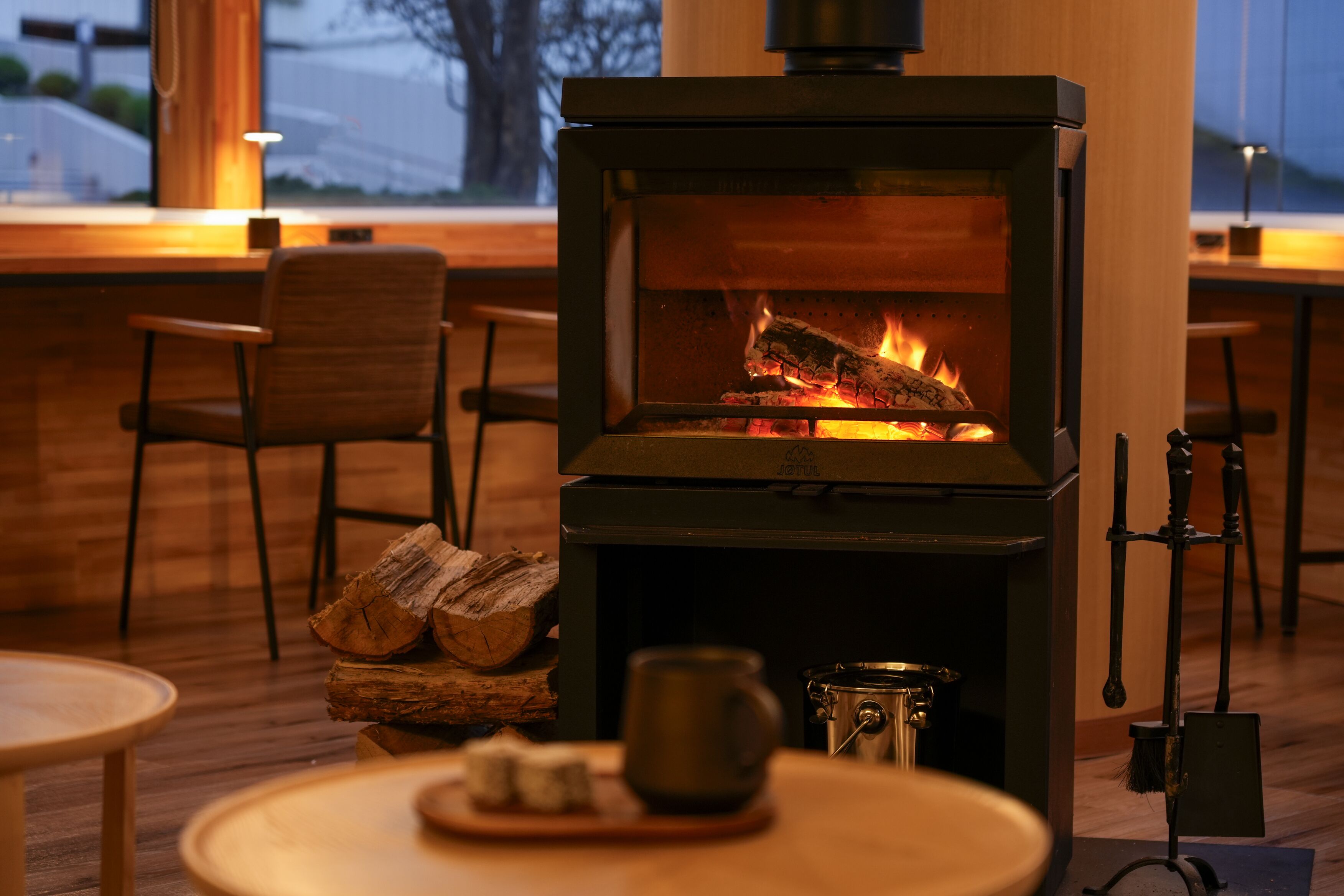 A wood-burning stove with logs, a metal pot, and a mug on a table in a cozy room with wooden furniture and a window.