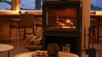 A wood-burning stove with logs, a metal pot, and a mug on a table in a cozy room with wooden furniture and a window.