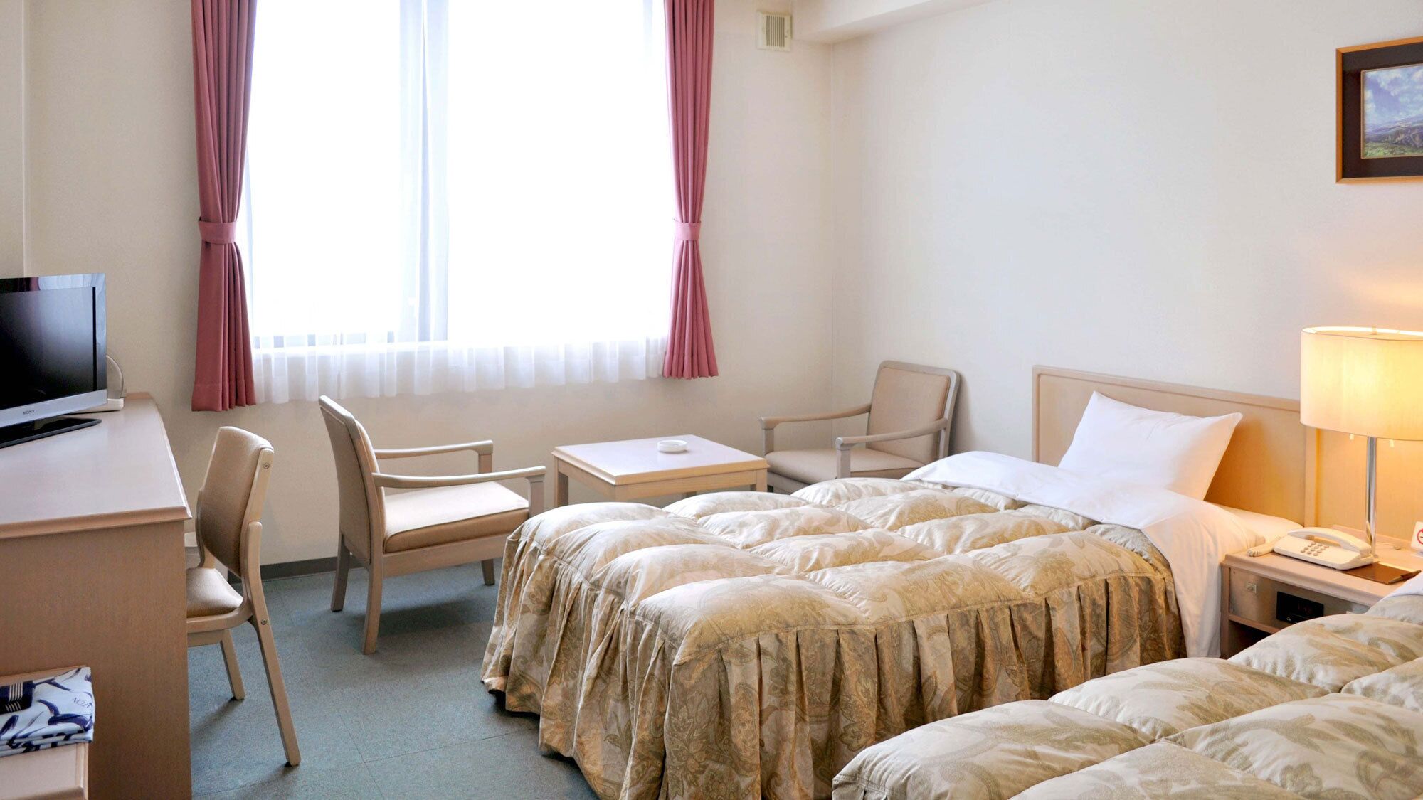 A hotel room with a bed, two chairs, a desk with a TV, a bedside table with a lamp, and a window with curtains.