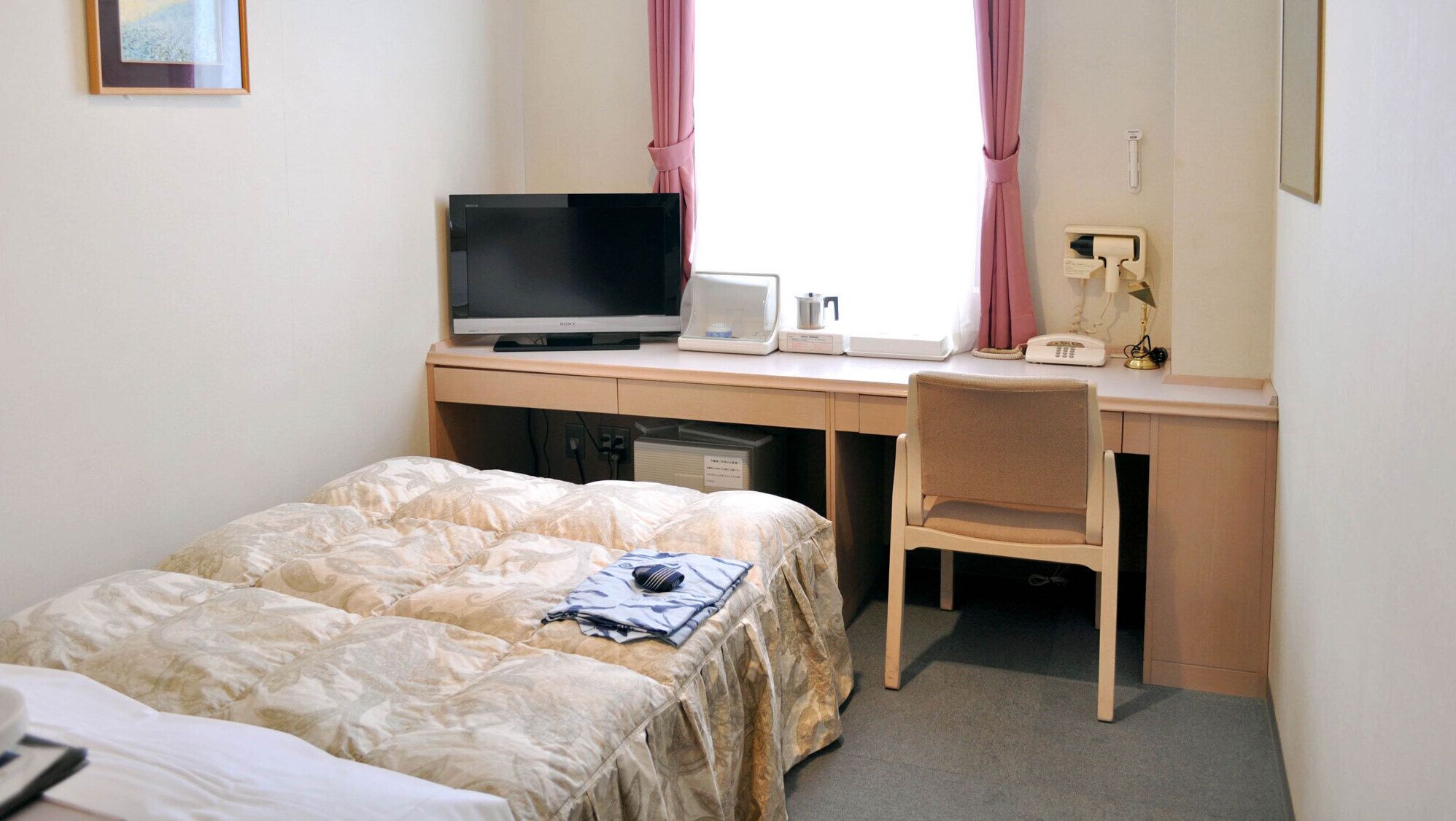 A hotel room with a bed, a desk with a television, a chair, and a window with curtains.