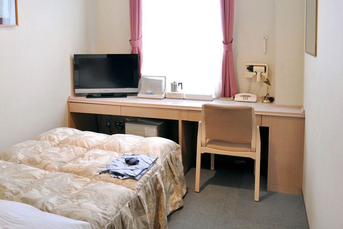 A hotel room with a bed, a desk with a television, a chair, and a window with curtains.