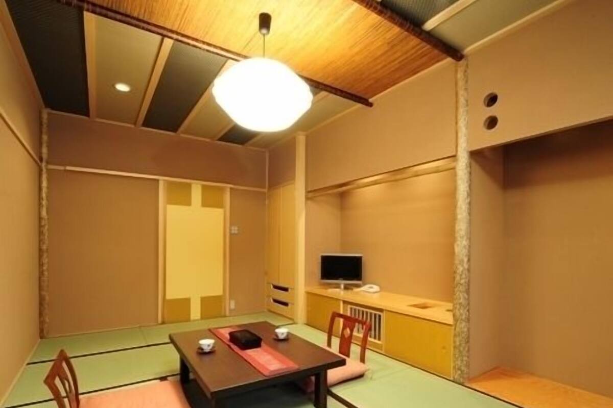 A tatami mat room with a low table, chairs, a television, and a ceiling light.