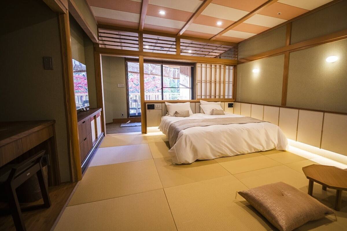 A traditional Japanese-style bedroom with tatami flooring, a bed with white bedding, a small wooden table, and a view of cherry blossoms through the window.