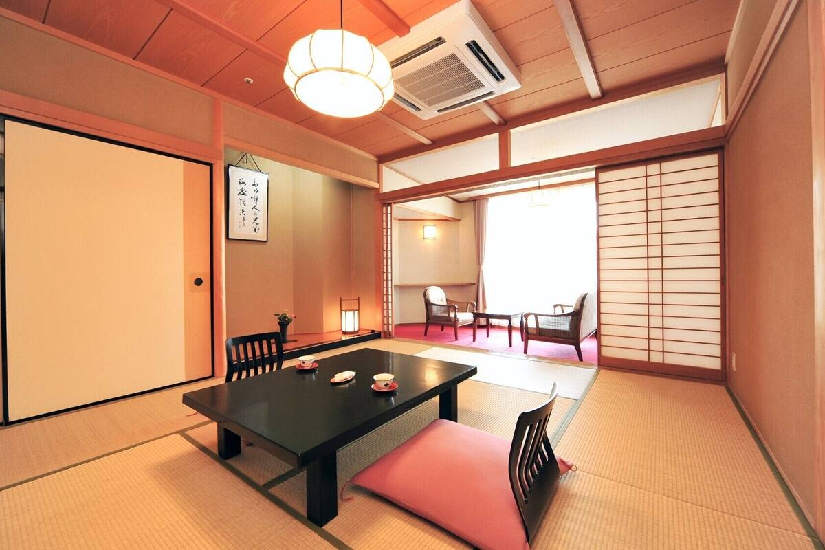A traditional Japanese room with tatami flooring, a low table with cushions, a sliding door, and a view of a garden through a shoji screen.
